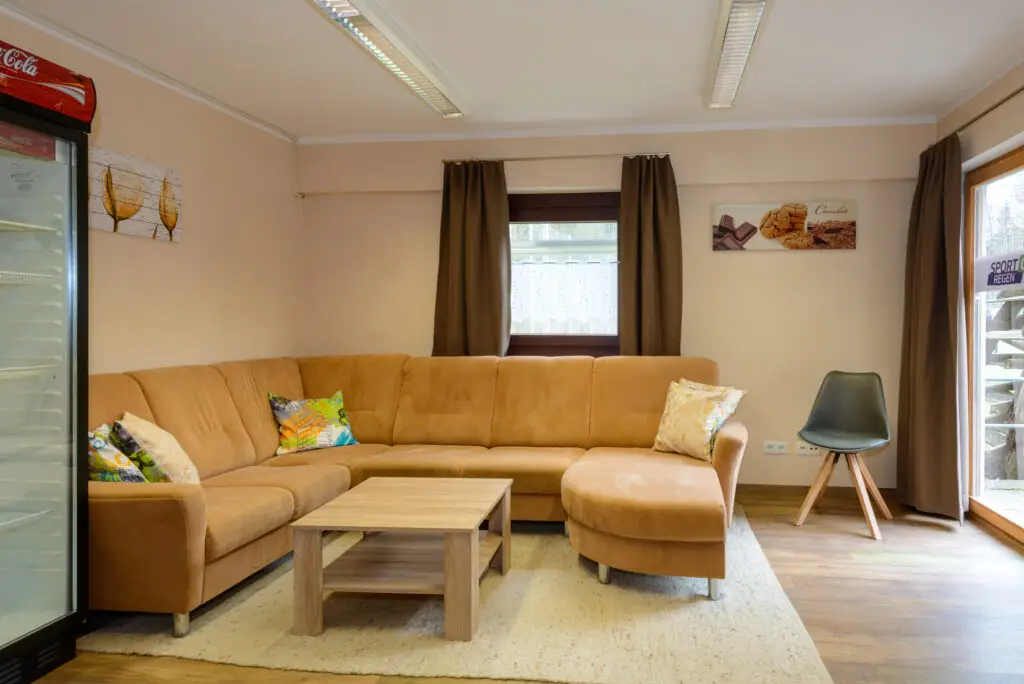 Wohnzimmer mit brauner Eckcouch, dekoriert mit bunten Kissen, Holzcouchtisch und grünem Stuhl. Links ein Kühlschrank, dahinter hängt ein Bild. Fenster im Hintergrund mit braunen Vorhängen und dahinter sichtbare Gartenlandschaft. Deckenleuchten sorgen für Beleuchtung.