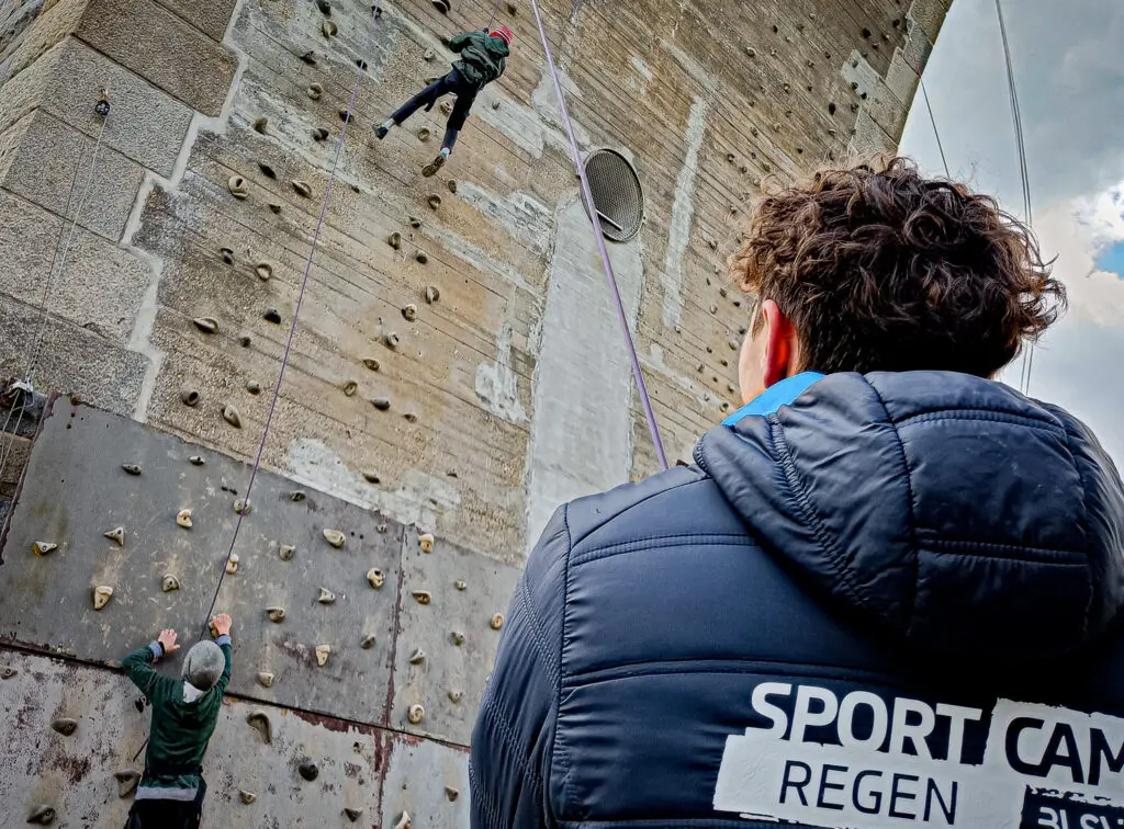 Eine Person klettert an einer Wand mit Klettergriffen, gesichert durch ein Seil. Ein Mann im Vordergrund, mit einer Jacke mit der Aufschrift "SPORT CAMP", beobachtet die Szene. Links unten klettert eine weitere Person die Wand hinauf.