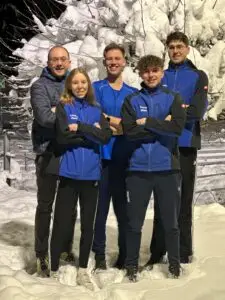 Fünf Personen stehen eng beieinander im Schnee. Sie tragen blaue Jacken und lächeln. Im Hintergrund sind Bäume mit Schnee bedeckt sichtbar. Die Szene wirkt wie ein gruppenfoto bei kaltem Wetter.